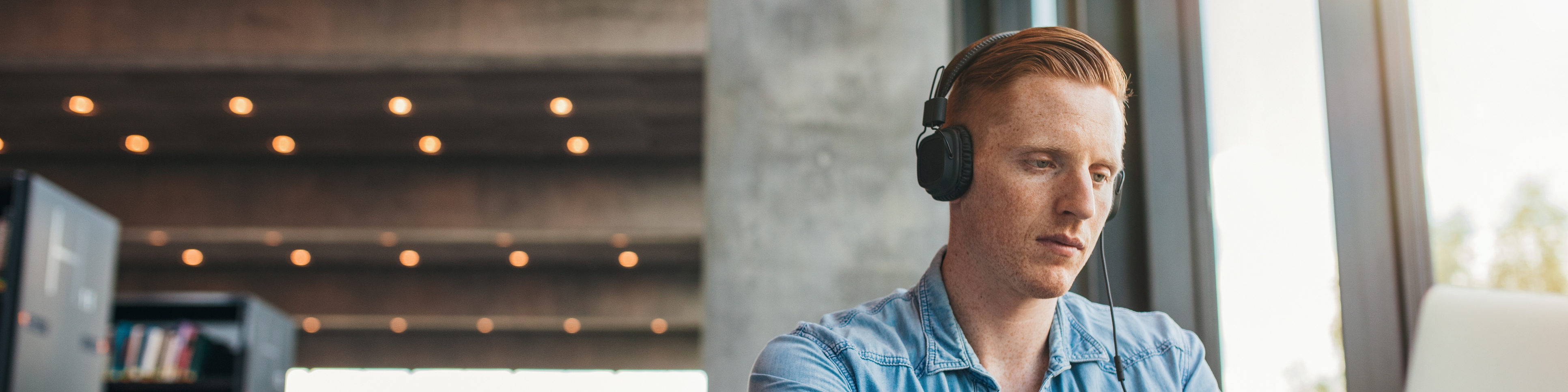 Junger Mann mit Kopfhörern sitzt an einem Laptop in einer Bibliothek